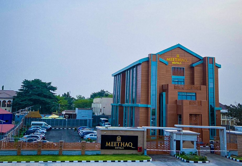 Photograph of the Meethaq Hotel exterior in Jabi, Abuja, showing its terracotta and teal facade with vertical glazing.